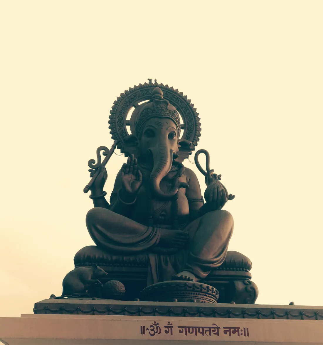 A dark Ganesha murti seated beneath a carved halo against a pale sky
